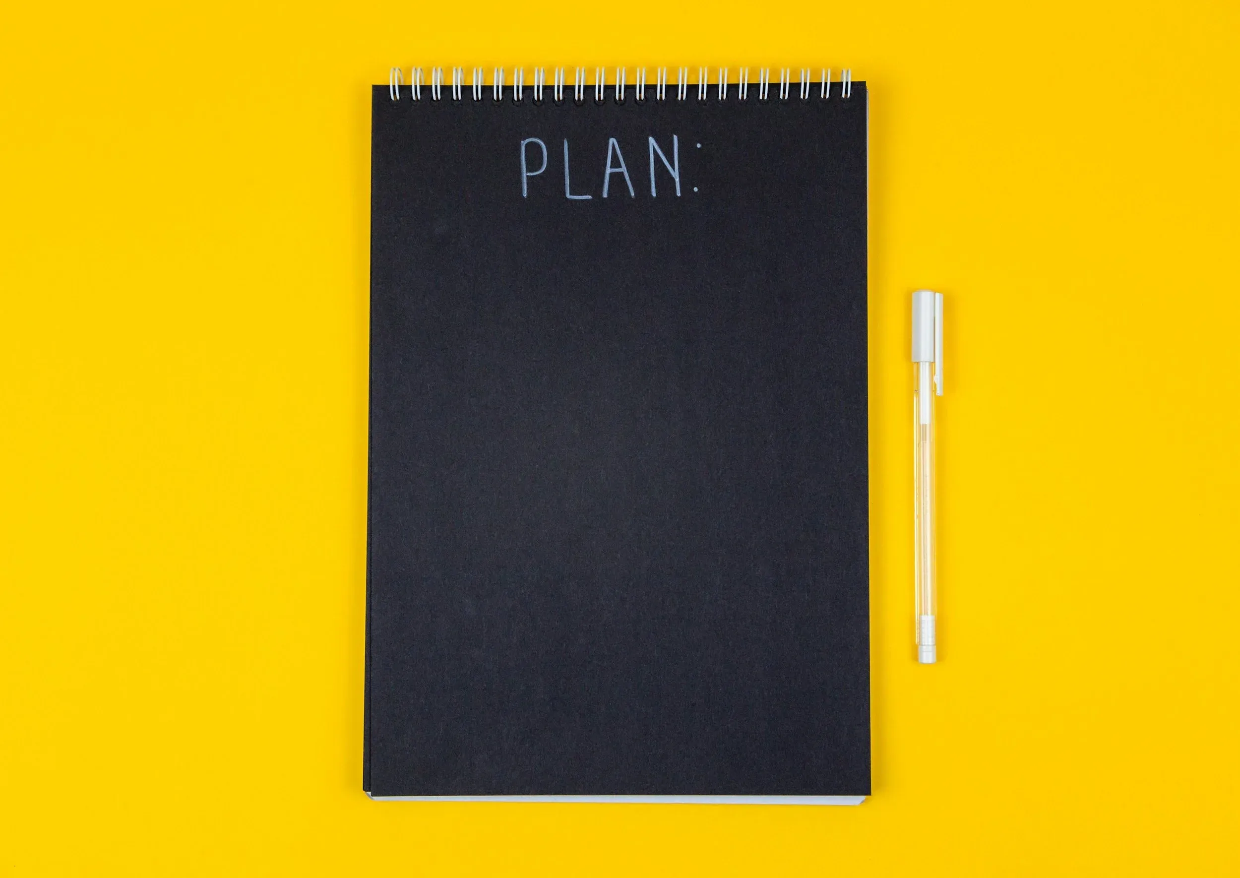  An image of a black spiral notebook with the word plan on it. There’s a pen next to it on a yellow background 