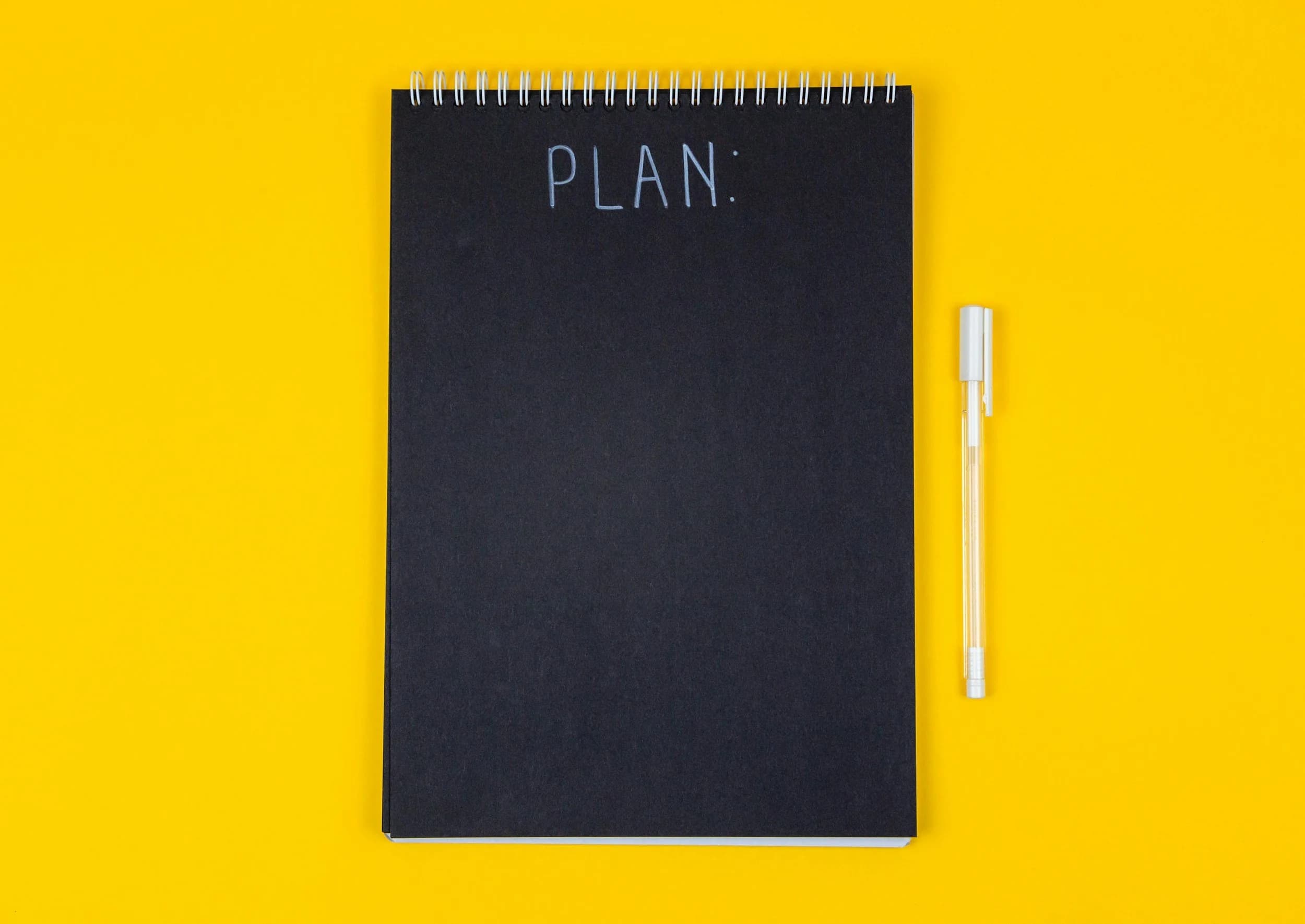  An image of a black spiral notebook with the word plan on it. There’s a pen next to it on a yellow background 